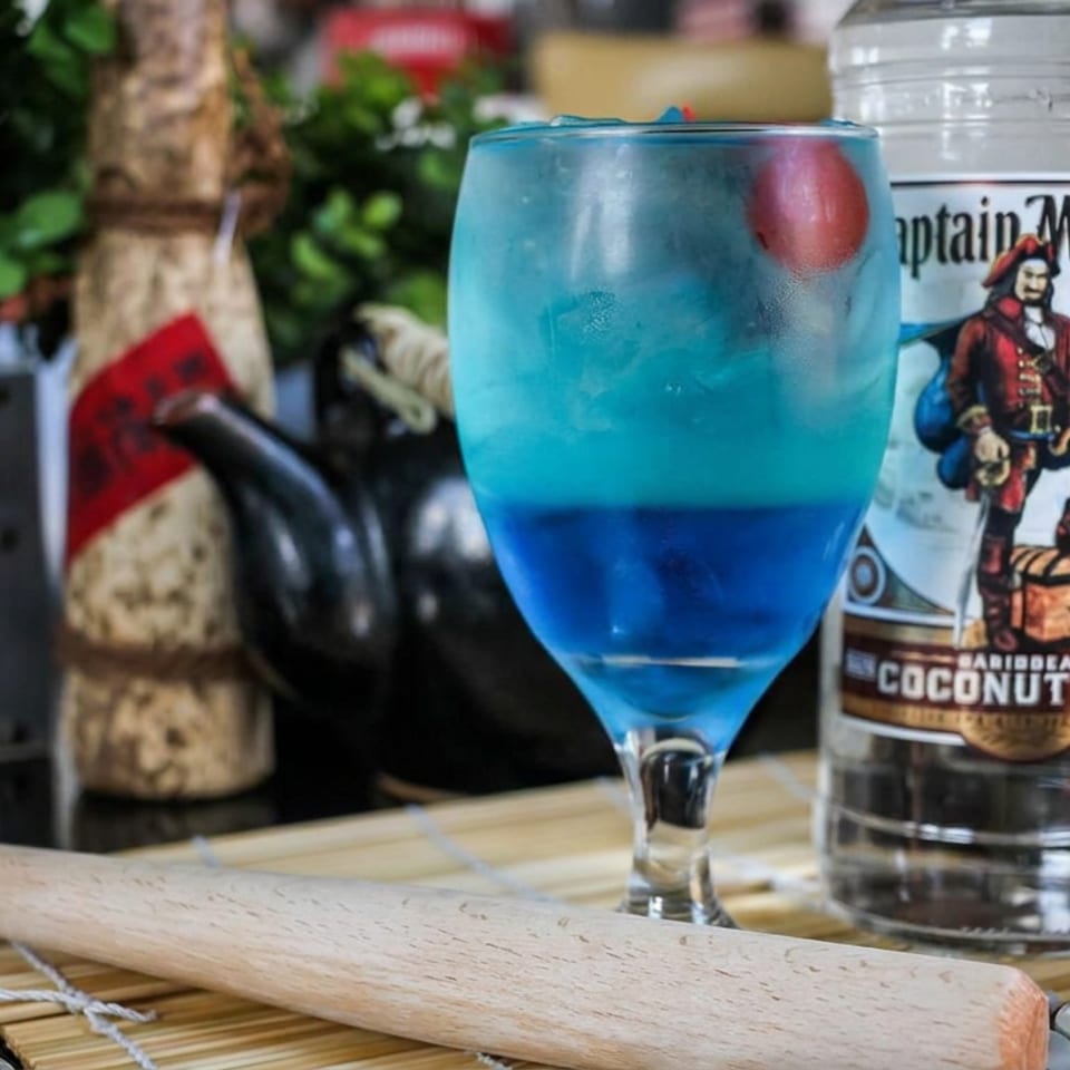 Blue cocktail with red fruit garnish beside a Captain Morgan Coconut Rum bottle and wooden rolling pin on a table.