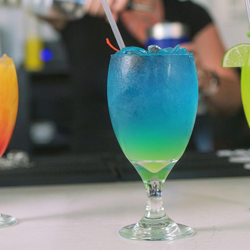 Three colorful cocktails—orange, blue, and lime green—served in stemmed glasses at a restaurant bar.