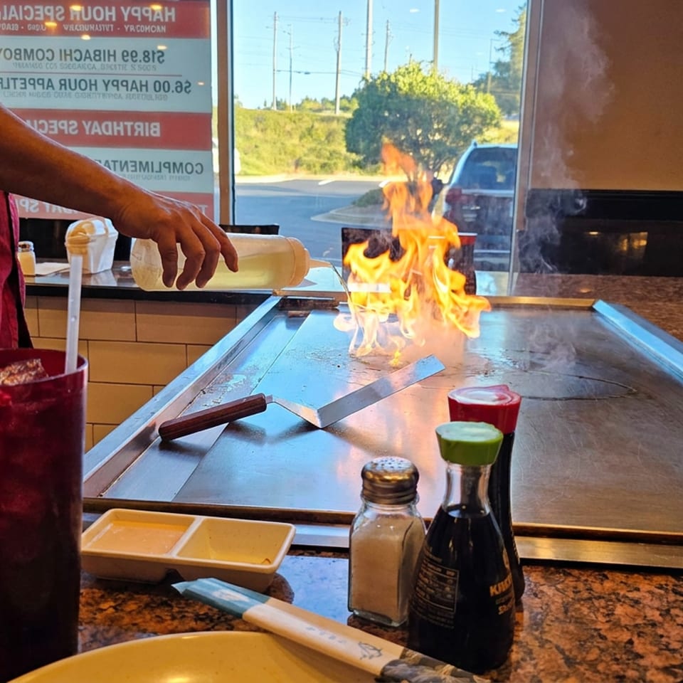 Restaurant chef pours sauce onto a hot teppanyaki grill, creating flames beside condiments and dining tables.