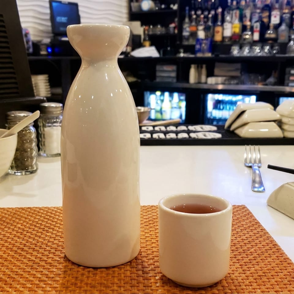White ceramic carafe and cup of tea on an orange woven placemat at a restaurant sushi counter.