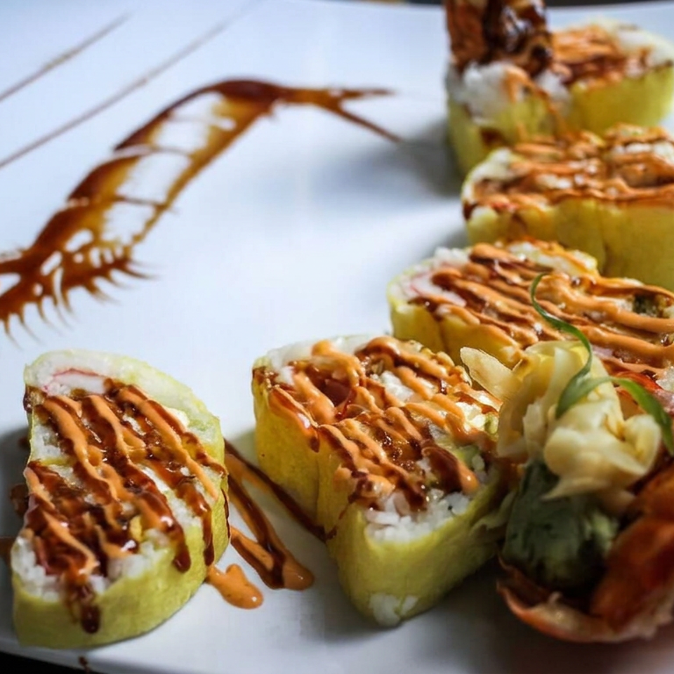 Sushi rolls drizzled with spicy sauce, accompanied by fried shrimp, vegetables, and decorative sauce on a white plate.