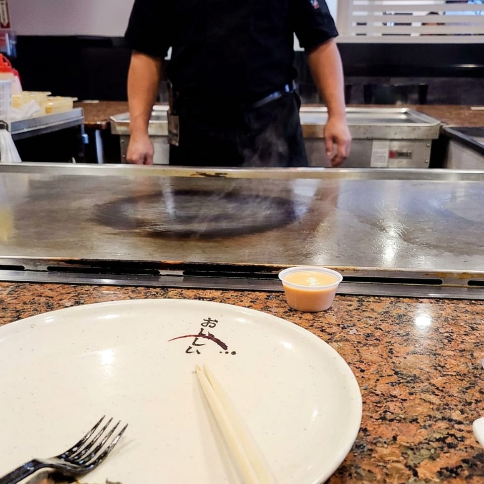 Chef stands behind a teppanyaki grill, with a plate, chopsticks, fork, and dipping sauces on the granite counter.