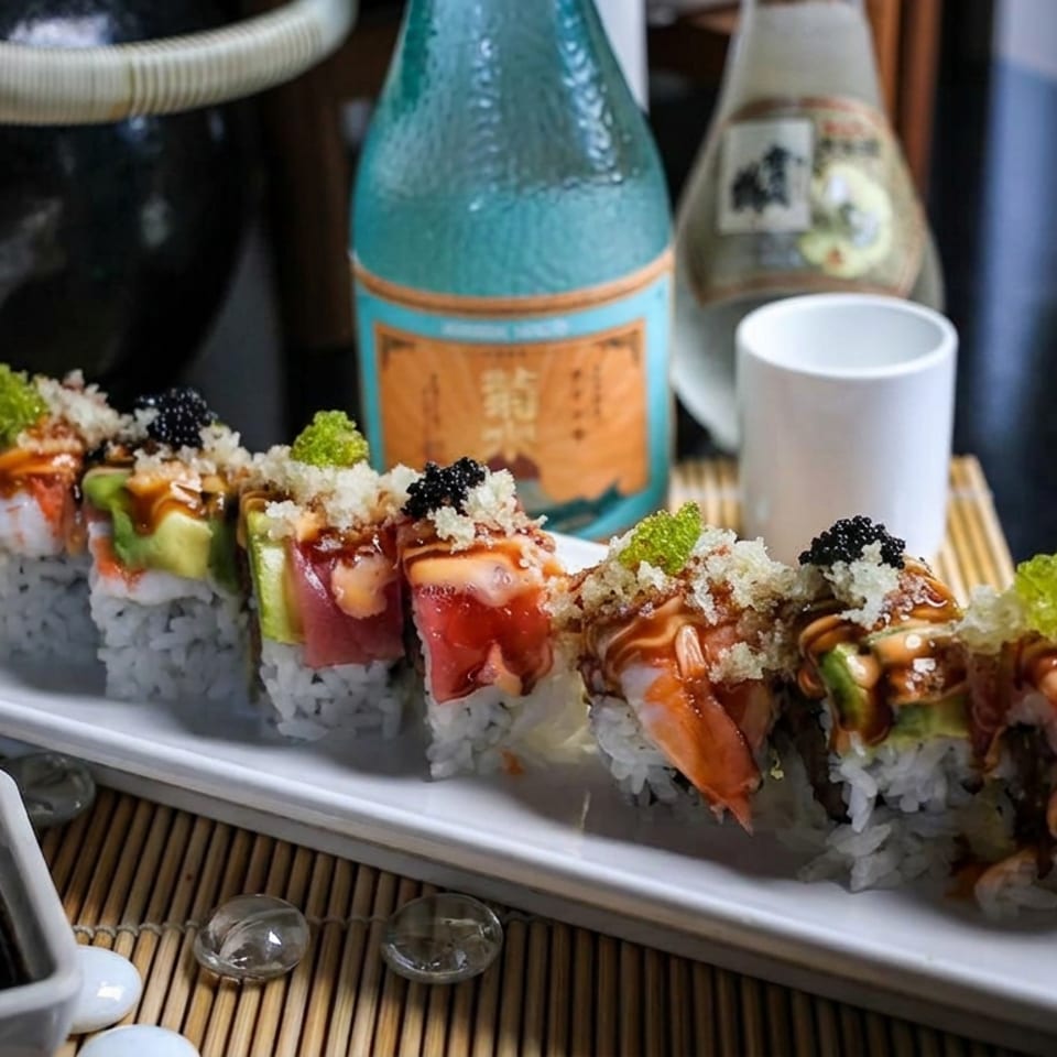 Colorful sushi rolls topped with fish, sauces, crispy crumbs, roe, and avocado garnish on a white platter with chopsticks and soy sauce.