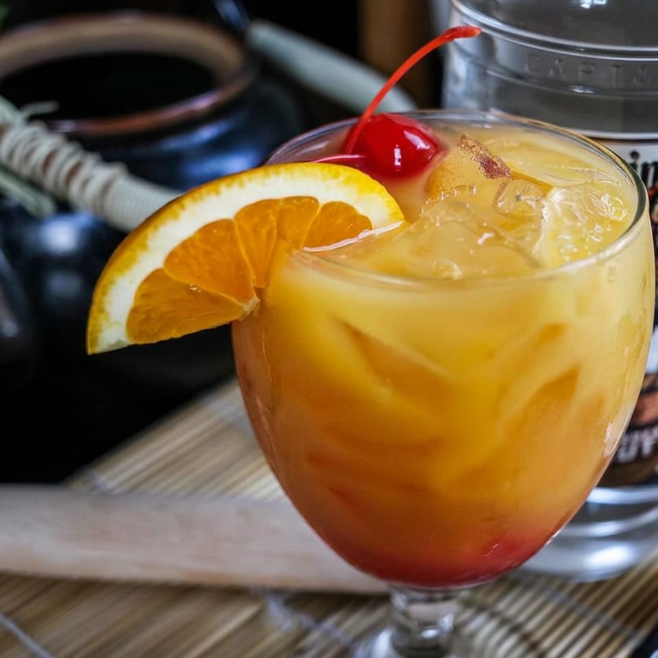 Orange cocktail with ice, cherry, and citrus wedge garnished beside a bottle and bar tools.