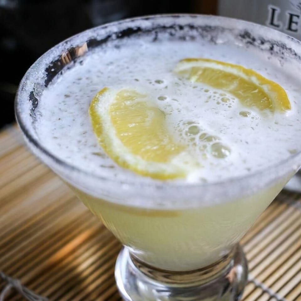 Frosted cocktail glass filled with a pale citrus drink, topped with lemon slices, on a bamboo placemat.