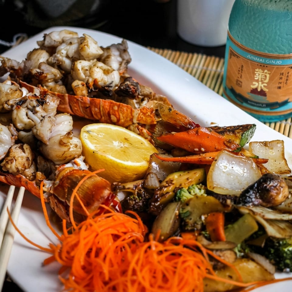 Grilled lobster tails and seafood served with roasted vegetables, lemon, dipping sauce, chopsticks, and a bottle of Japanese sake.