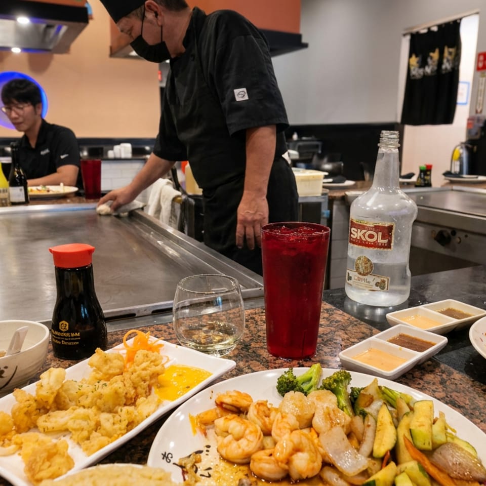 Teppanyaki chefs prepare food behind a hot grill surrounded by plates of shrimp, vegetables, tempura, sauces, and drinks.