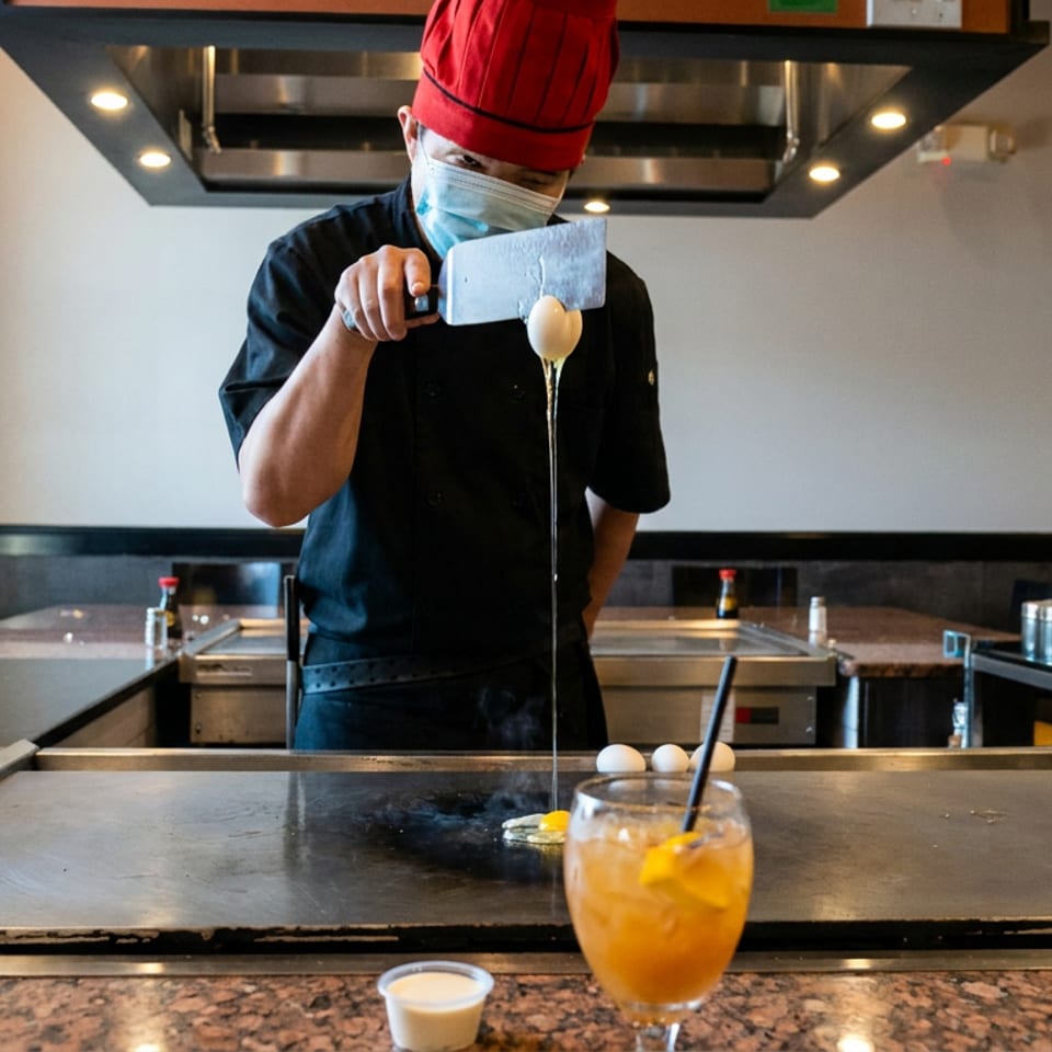 Masked chef in a red hat cracks an egg over a teppanyaki grill while an orange drink sits in the foreground.